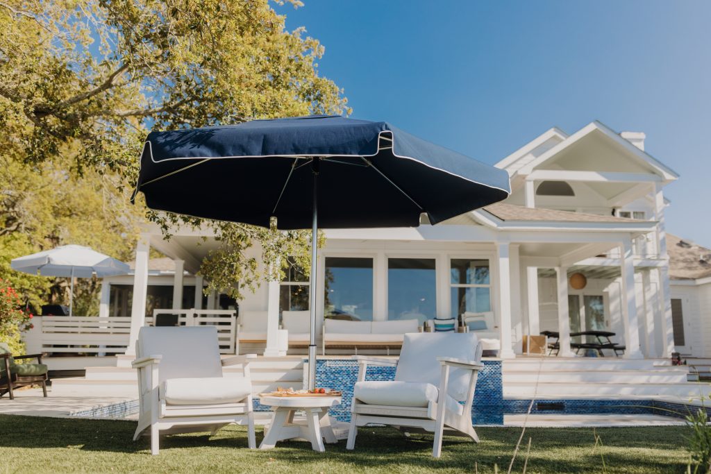 A luxurious backyard scene featuring a navy outdoor umbrella with white piping, shading two cushioned white patio chairs and a small round table. The table holds a wooden tray with snacks, positioned on green grass in front of a modern white home with large windows, a shaded porch, and a blue-tiled pool. The bright, sunny weather and surrounding trees complete the relaxed coastal atmosphere.